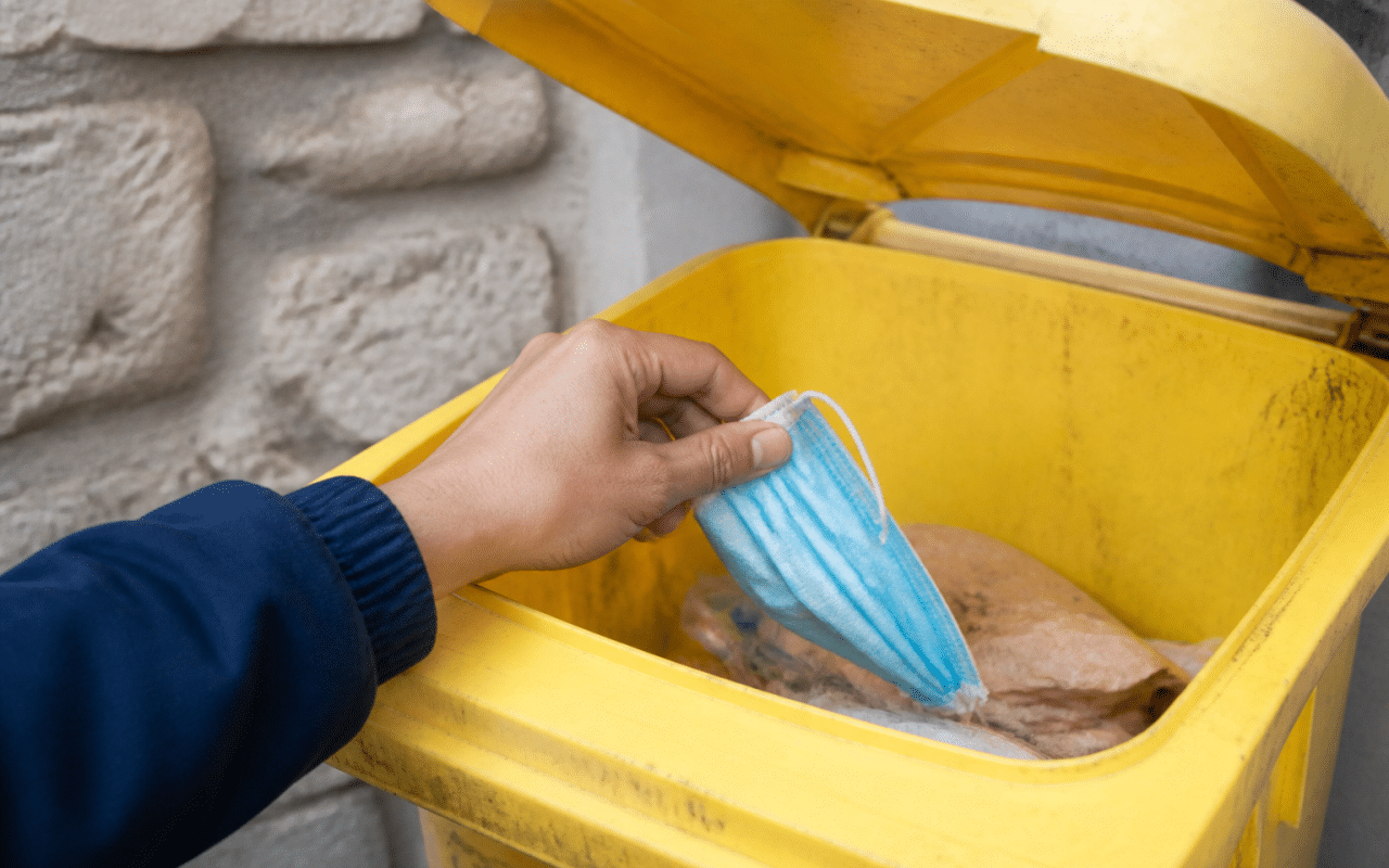Main tenant masque bleu dans poubelle jaune ouverte